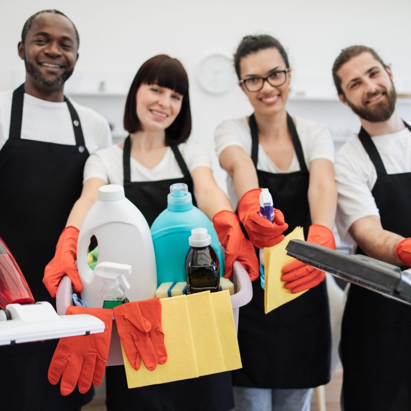 Professional cleaning service. Portrait of happy multiethnic janitors holding cleaning equipment against background of light modern kitchen. Cleaning team, group of four multiracial people in black aprons and red gloves.